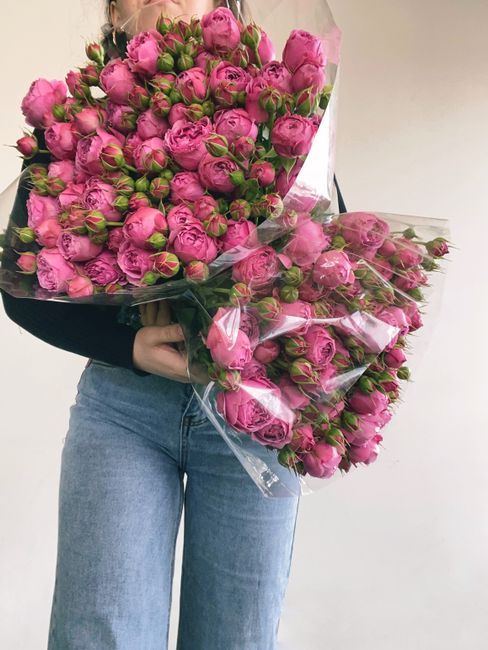 A person is holding a large, tightly clustered bouquet of vibrant pink roses with green stems and buds, wrapped in clear plastic. The flowers are fresh with smooth, velvety petals and lush foliage, ar