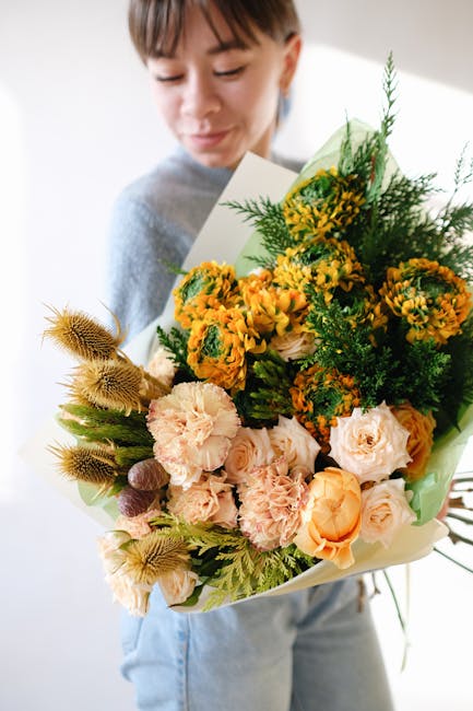 A woman holding a large floral bouquet featuring a mix of yellow and orange flowers, including spray chrysanthemums and roses, complemented by green fern and foliage accents, wrapped in light green an