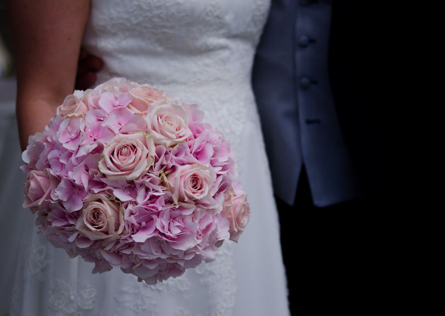 A close-up of a bridal bouquet featuring soft pink roses, meticulously arranged with pink hydrangeas, held by a bride in a white lace dress. The bouquet appears fresh, with velvety petals and sturdy g