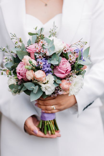 A person dressed in a white blazer holds a bouquet of fresh flowers with both hands. The bouquet features pink roses, white lisianthus, small purple and pink filler flowers, and green eucalyptus leave